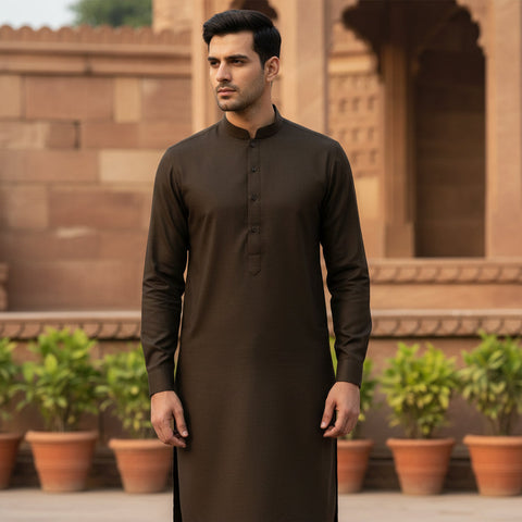 Man wearing a brown traditional outfit standing in front of a brick wall with plants.