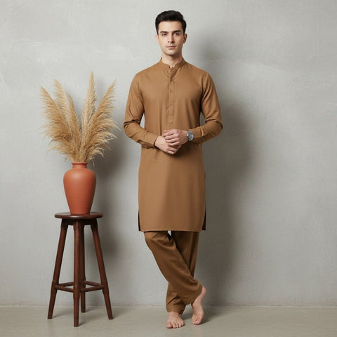 Man wearing a brown kurta standing next to a wooden stool with a vase and dried plants against a plain wall.