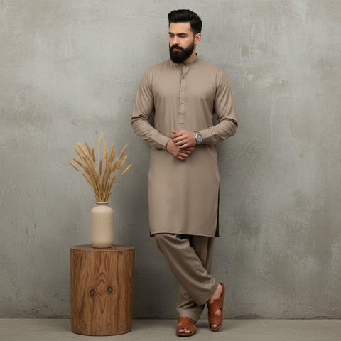 Man wearing a beige kurta with pants standing next to a wooden stool with a vase against a gray wall.