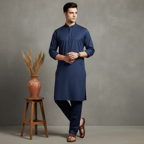 Man wearing a blue kurta standing next to a wooden stool with a vase on it against a gray background