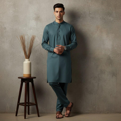 Man wearing a teal kurta standing next to a wooden stool with a decorative vase against a gray wall.