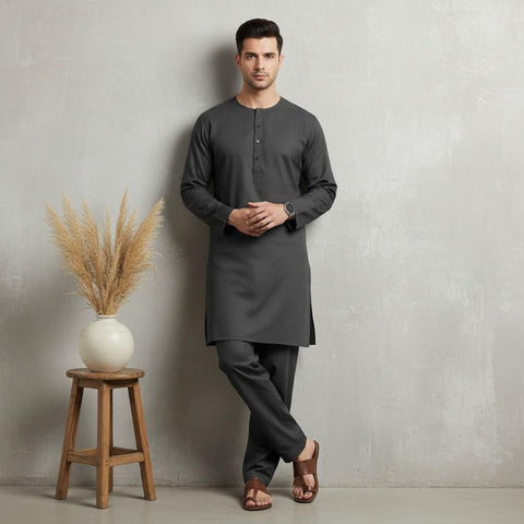 Man wearing a dark gray kurta standing against a plain wall with a decorative plant on a stool.