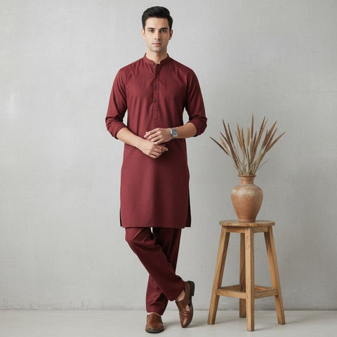 Man wearing a maroon kurta with pants standing next to a wooden stool with a vase.
