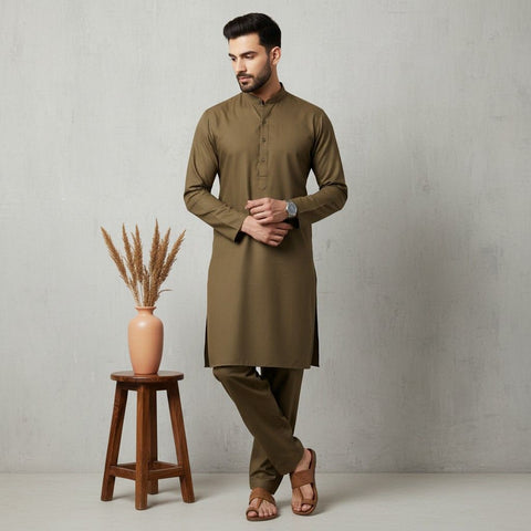 Man wearing a brown kurta standing next to a wooden stool with a vase containing dried plants on a plain background