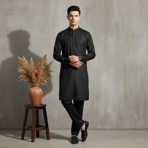 Man wearing a black kurta standing next to a wooden stool with a vase and dried plants against a gray wall.