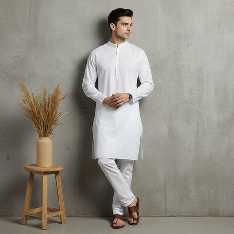 Man wearing a white kurta standing next to a wooden stool with a vase of dried plants against a gray wall.