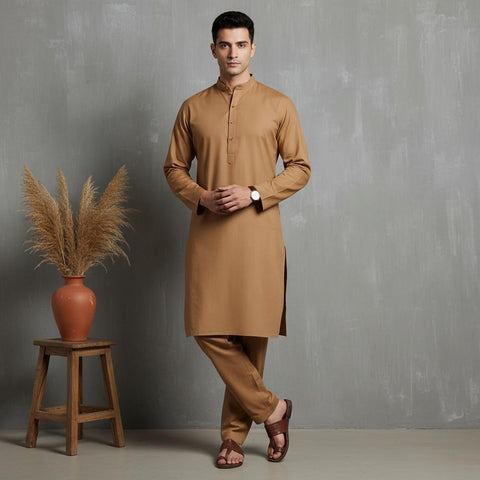 Man wearing a brown kurta standing next to a wooden stool with a vase and dried plants against a gray wall.