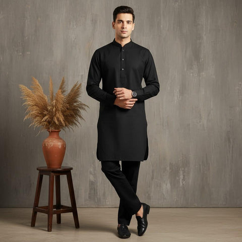 Man wearing a black kurta standing next to a wooden stool with a vase of dried plants against a gray wall.