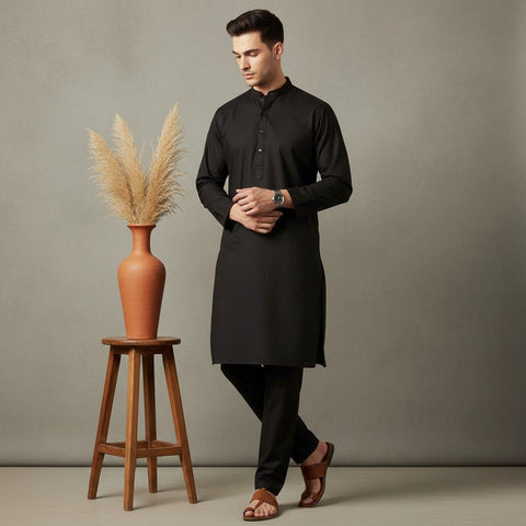 Man wearing a black kurta standing next to a wooden stool with a vase and dried plants against a plain background