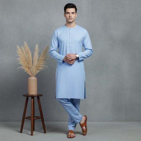 Man wearing a light blue kurta standing next to a wooden stool with a vase containing dried plants on a gray background