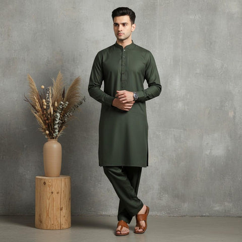 Man wearing a green kurta standing next to a wooden stool with a vase of dried plants against a gray wall.