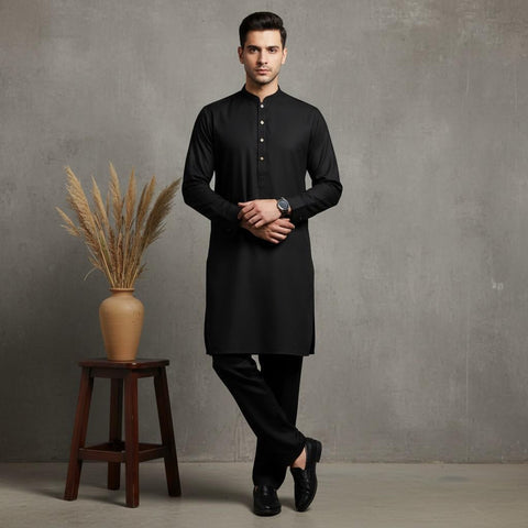 Man wearing a black kurta standing next to a wooden stool with a vase containing dried plants against a gray wall.