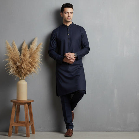 Man wearing a navy kurta standing next to a wooden stool with a vase of dried plants against a gray wall.