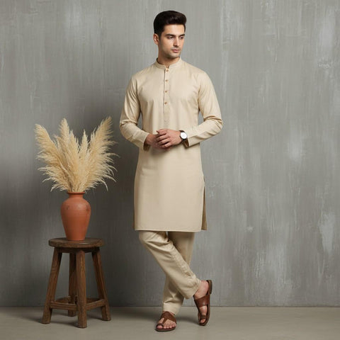 Man wearing a beige kurta standing next to a wooden stool with a vase of dried plants against a gray background