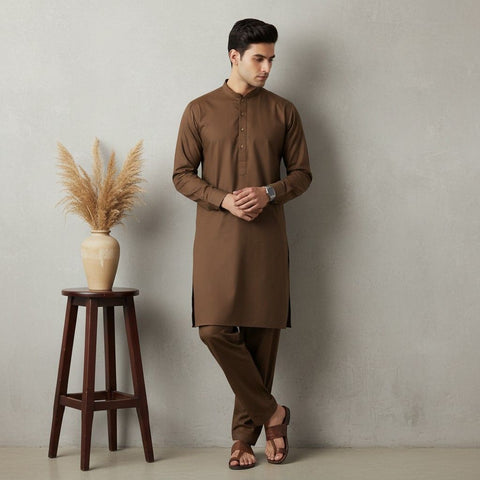 Man wearing a brown kurta standing next to a wooden stool with a vase containing dried plants against a plain wall.