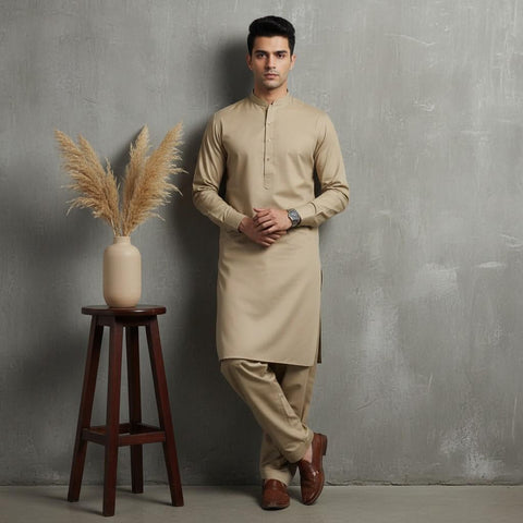 Man wearing a beige kurta standing next to a wooden stool with a vase and dried plants against a gray wall.