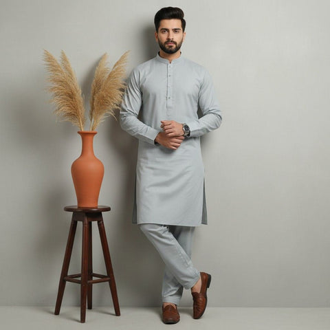 Man in a light gray kurta standing next to a wooden stool with an orange vase and dried plants on a plain background