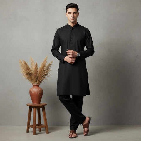 Man wearing a black kurta standing next to a wooden stool with a vase and dried plants against a gray background