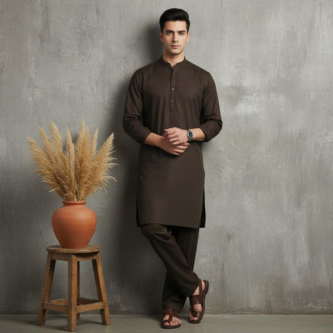 Man wearing a brown kurta standing next to a wooden stool with a vase of dried plants against a gray wall.