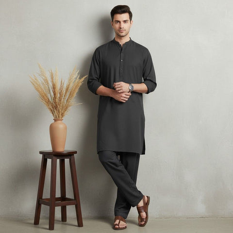Man wearing a black kurta standing next to a wooden stool with a vase of dried plants against a plain wall.