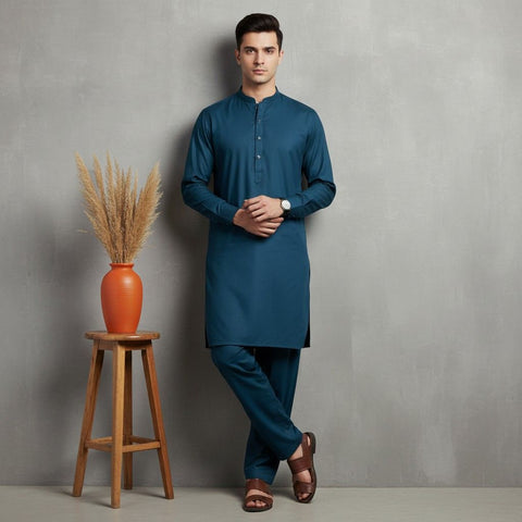 Man wearing a blue kurta standing next to a wooden stool with an orange vase and dried plants against a gray wall.