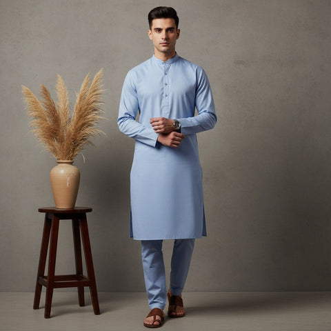 Man wearing a light blue kurta standing next to a wooden stool with a vase and dried plants against a gray background.