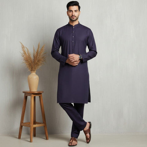 Man wearing a dark purple kurta with black pants standing next to a wooden stool with a vase containing dried plants on a light gray background.