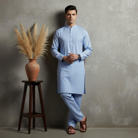 Man wearing a light blue kurta standing next to a wooden stool with a vase and dried plants against a gray wall.