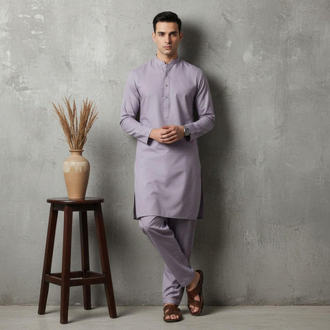 Man wearing a light purple kurta standing next to a wooden stool with a vase against a gray wall.