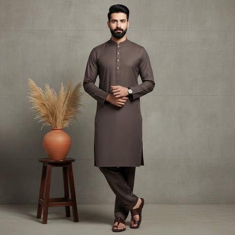 Man wearing a brown kurta standing next to a wooden stool with a vase and dried plants against a gray wall.