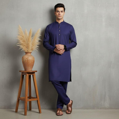 Man wearing a purple kurta standing next to a wooden stool with a vase and dried plants against a gray wall.