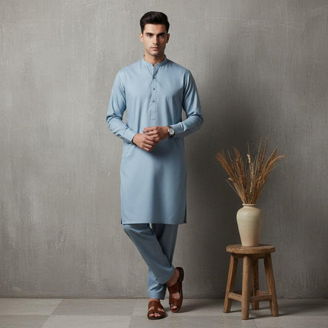 Man wearing a light blue kurta standing next to a wooden stool with a vase against a gray wall.