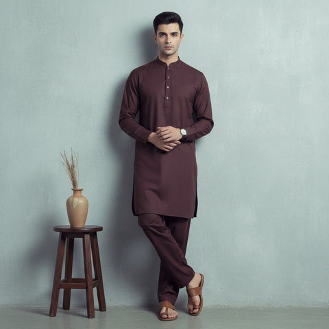 Man wearing a brown kurta standing next to a wooden stool with a vase against a gray wall.