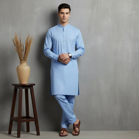 Man wearing a light blue kurta standing next to a wooden stool with a vase against a gray wall.