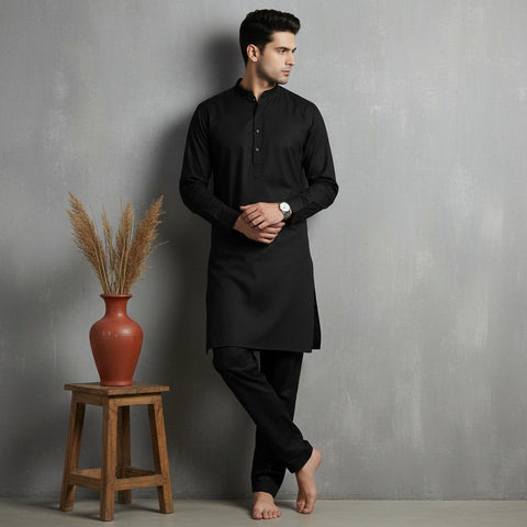 Man wearing a black kurta standing next to a wooden stool with a vase against a gray wall.