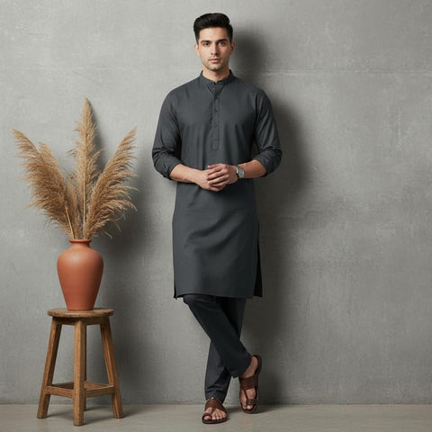 Man wearing a dark gray kurta standing next to a wooden stool with a vase and plant against a gray wall.