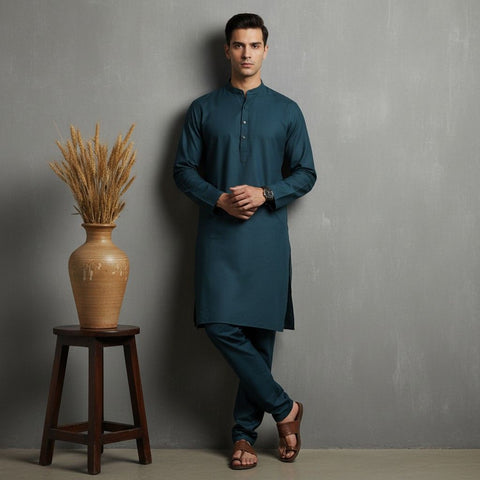 Man wearing a teal kurta standing next to a wooden stool with a vase containing dried plants against a gray wall.