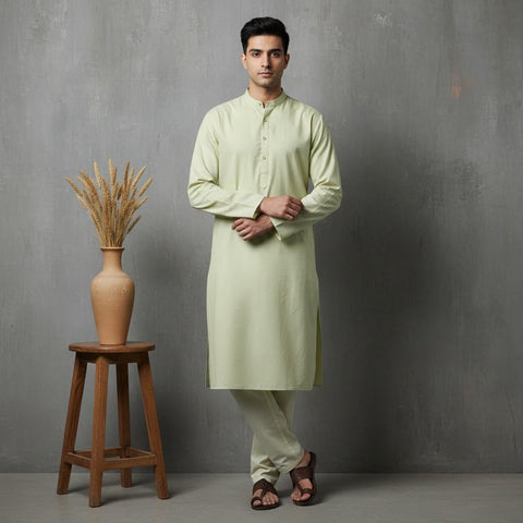 Man wearing a light green kurta standing next to a wooden stool with a vase on it against a gray wall.